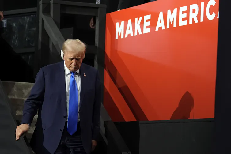 Donald Trump wearing a dark suit and a bandage on his right ear walking past a make america great again sign