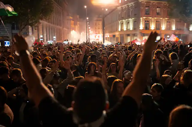 A crowd is seen through the outstretched arms of someone celebrating in a city square at night.