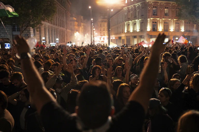 A crowd is seen through the outstretched arms of someone celebrating in a city square at night.