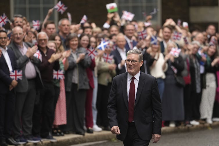 A man with glasses in a dark suit walks beside a crowd of people cheering behind barricades and waving the Union Jack.