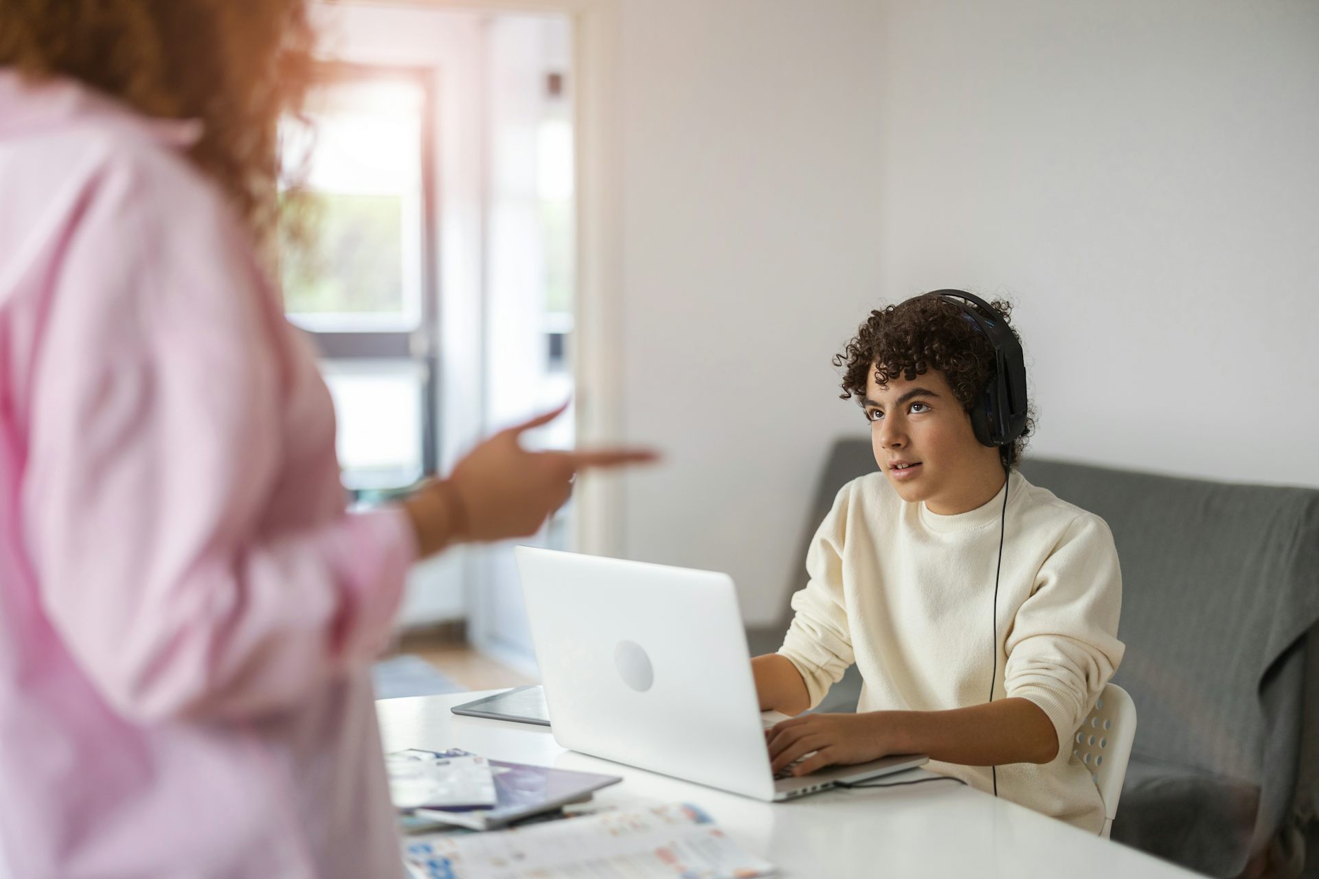 A teenage boy on a laptop with headphones, his mother talking to him.