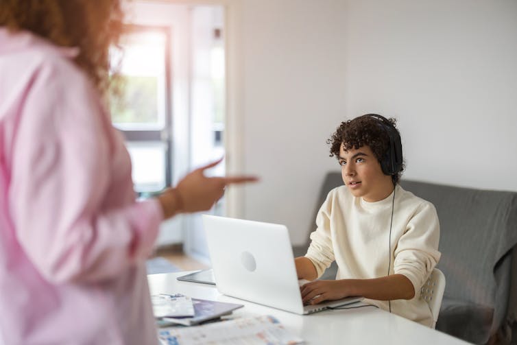 A teenage boy on a laptop with headphones, his mother talking to him.
