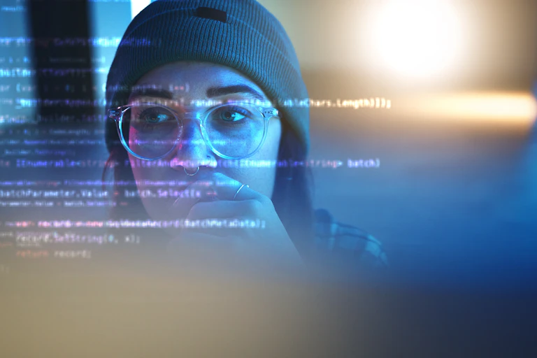 A woman in a beanie and glasses looks at a transparent screen with code on it.