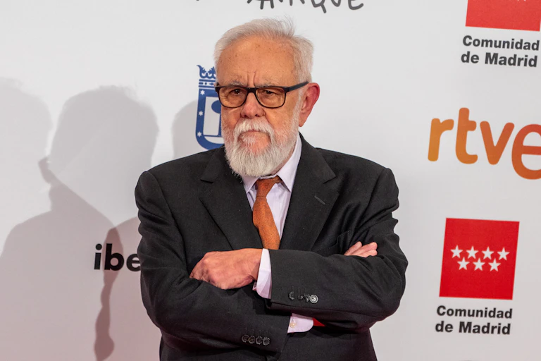 Un hombre con gafas y pelo y barba blanca, y de traje, cruza los brazos y mira a cámara.