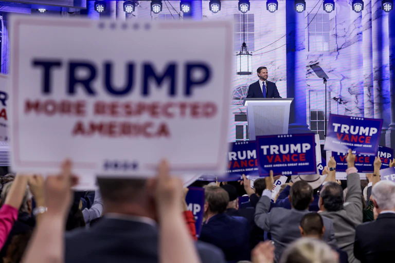 JD Vance speaks at a podium at the Republican National Convention as supporters wavew signs reading Trump/Vance. In the foreground is a placard reading Trump: More respected America.