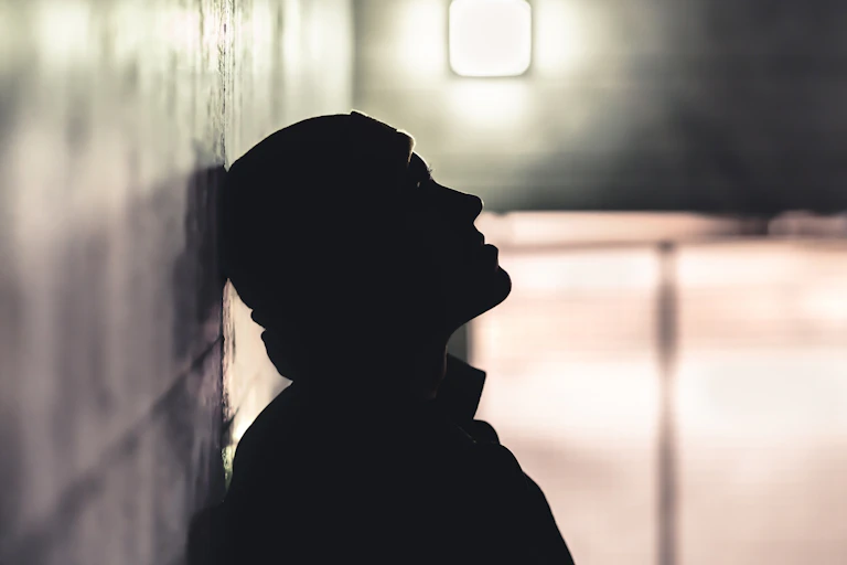 Black and white silhouette of a man resting his head against an external wall, a light glows behind him