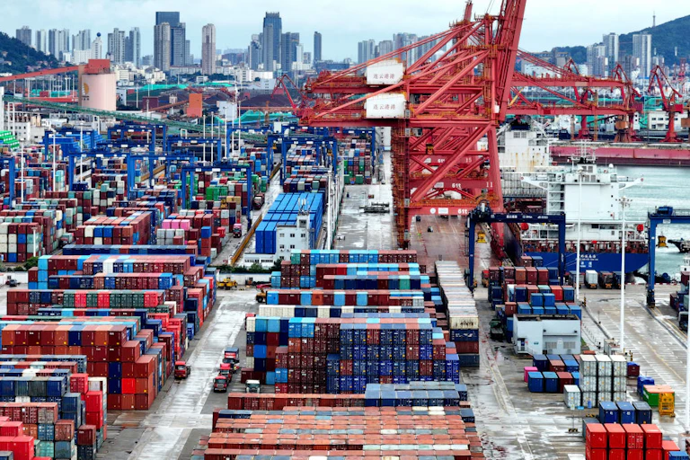 Stacks of shipping containers and some cranes in the foreground and tall city buildings in the distance