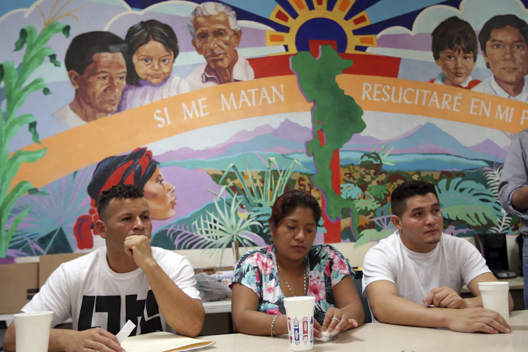Two men and a woman sit at a table. In the background is a large poster with words in Spanish: 'Si me matan, resucitare in mi pueblo,' or 'If they kill me, I will be reborn.'