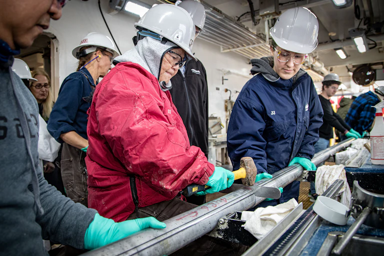 Technicians support a long metal tube, with one pounding a slicer in with a mallet