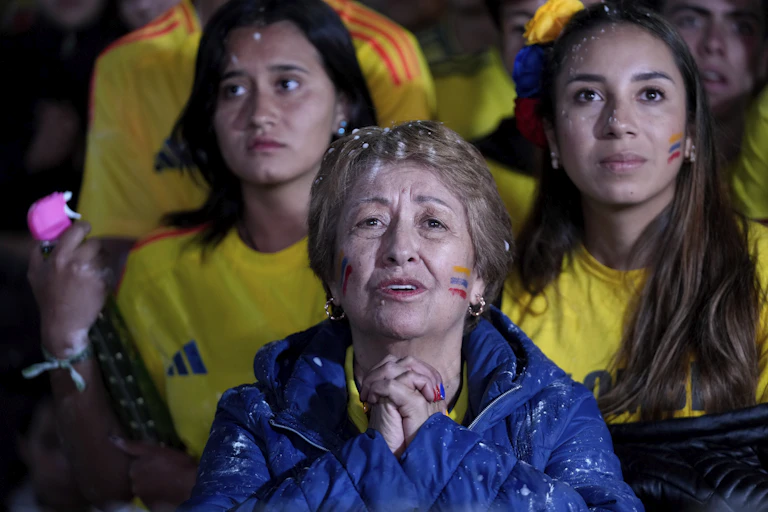 An elderly woman in a blue jacket sits with her hands clasped in front of her