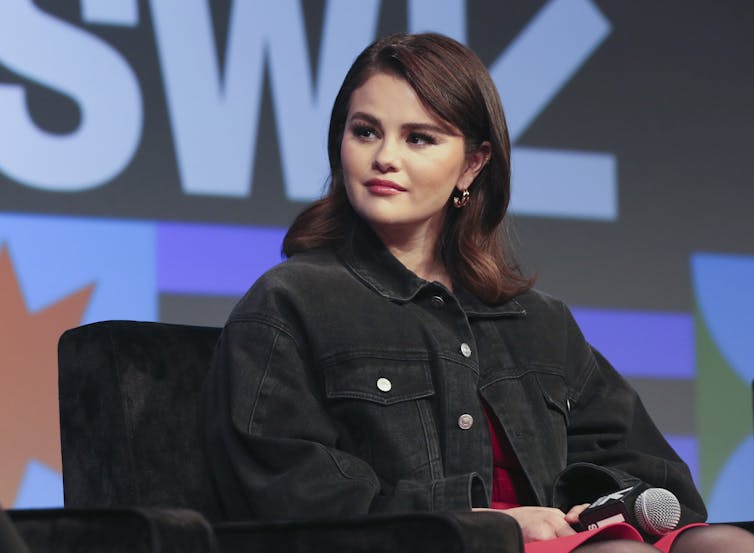 A young woman with shoulder-length hair in a black denim jacket sits in a chair in front of a large screen