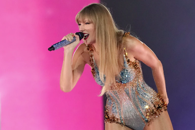 A white blonde woman ina sparkling body suit smiles while singing into a microphone