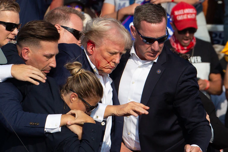 a man bleeding from his ear is surrounded by people wearing blue blazers and sunglasses