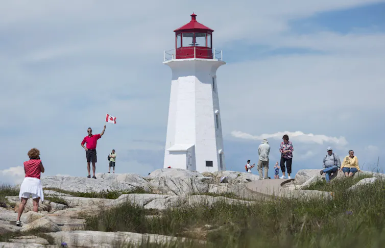 A woman in a red shirt takes a photo of a man waving a Canadian flag in front of a lighthouse