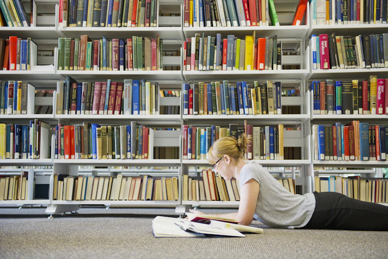 A young woman lies on the floor of a library. There are five rows of books behind her.