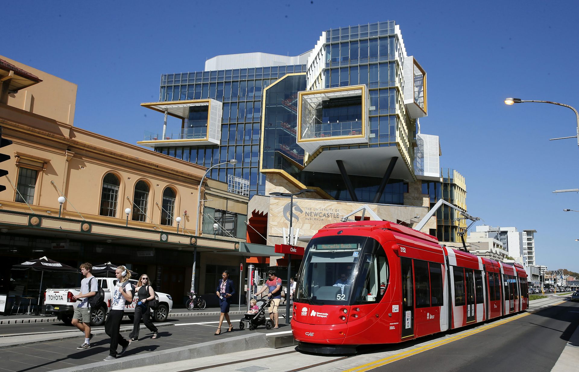 A tram is topped outside low-rise buildings on a street. A University of Newcastle building stands in the background.