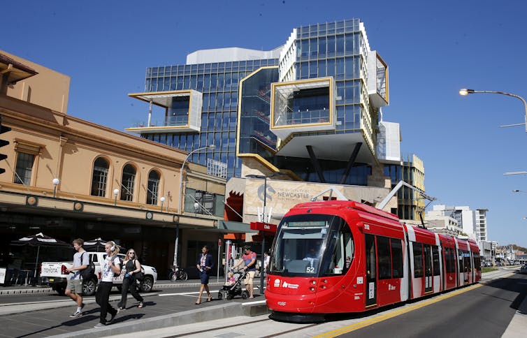 A tram is topped outside low-rise buildings on a street. A University of Newcastle building stands in the background.