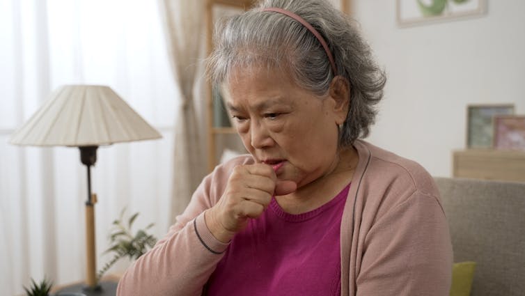 Woman of Asian heritage sitting in living room coughing, hand to mouth