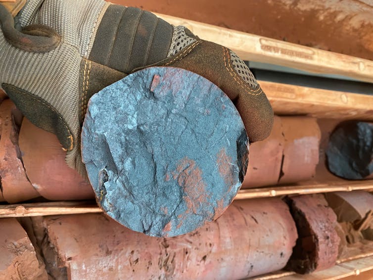 A gloved hand holding a round steely blue rock on a backdrop of reddish rocks.