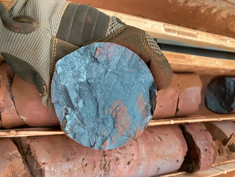 A gloved hand holding a round steely blue rock on a backdrop of reddish rocks.