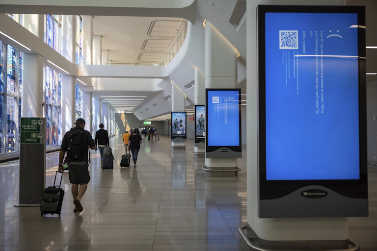 Several people pulling rolling luggage walk down a long hall past large display screens