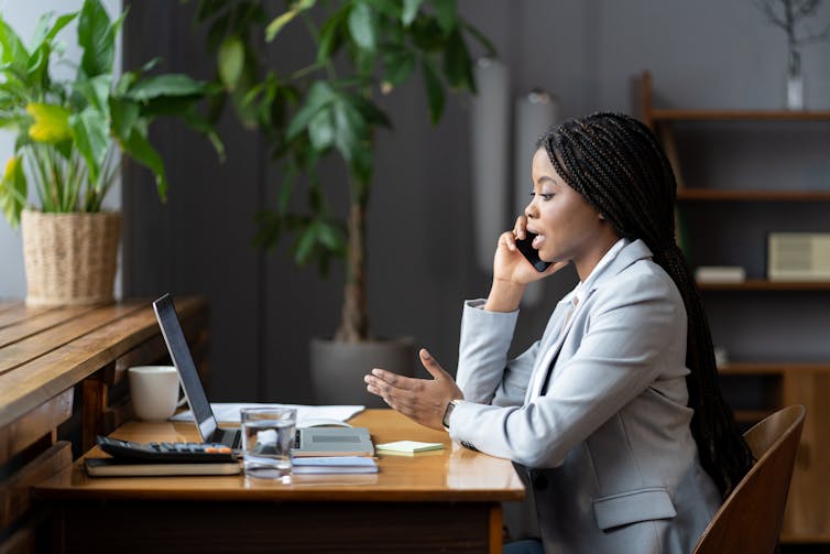 Side view of a young woman in a grey suit talking on the phone while looking at an open laptop