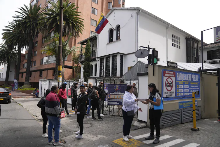 People gather in front of a white building flying a Venezuelan flag.