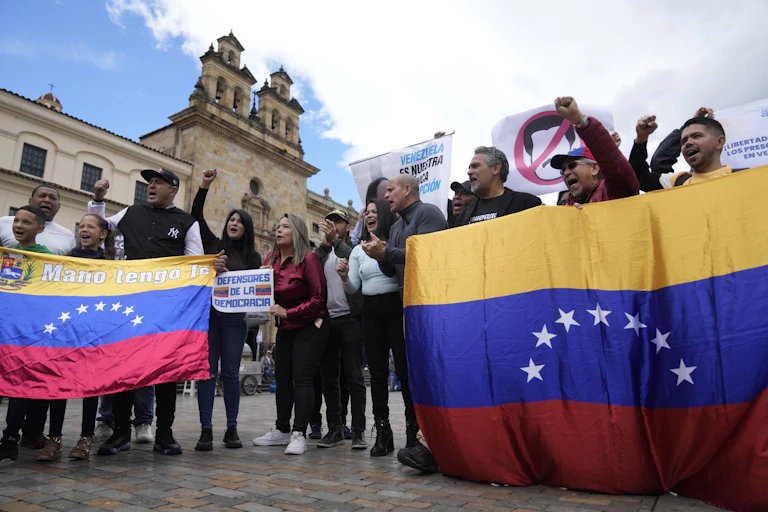People chant at a protest with their fists raised. They carry large Venezuelan flags.
