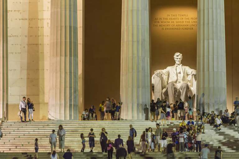A few dozen people stand and sit on white steps leading to a marble statue of a seated man behind tall pillars.