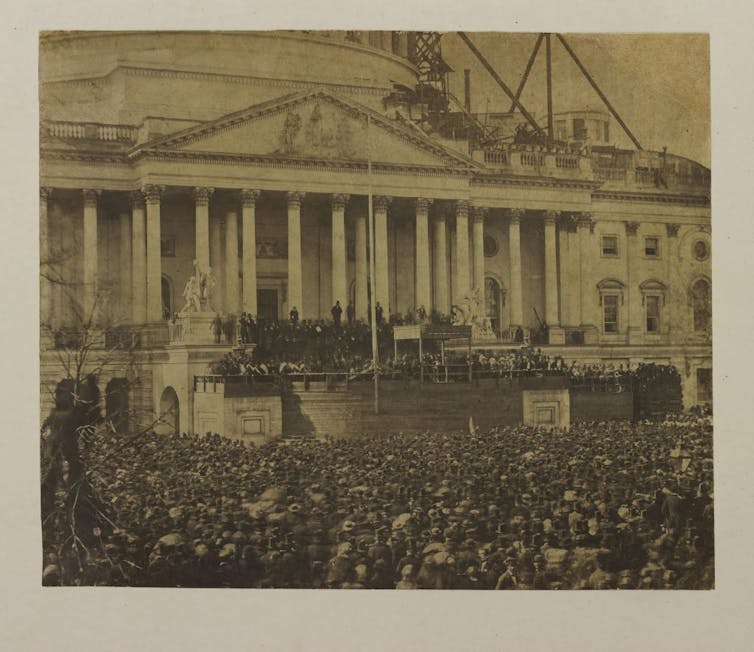 A sepia-toned image of the White House, with part of the building under repair, and a large crowd outside, mostly wearing black.