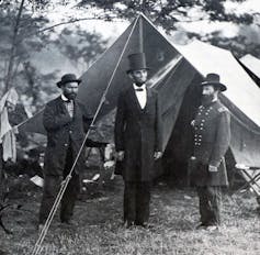 Three men wearing dark coats and hats, one of whom is much taller, stand outside a simple tent.