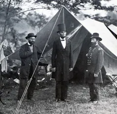 Three men wearing dark coats and hats, one of whom is much taller, stand outside a simple tent.
