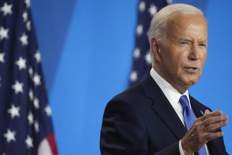 A white haired man in blue blazer standing in front of two flags.