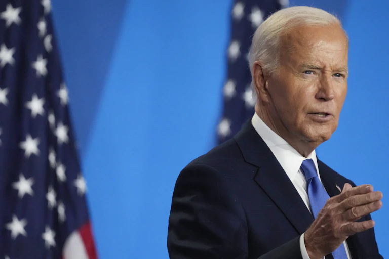 A white haired man in blue blazer standing in front of two flags.