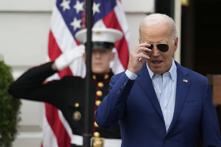 An older man with grey thinning hair wearing a stylish navy jacket removes his aviator sunglasses. a marine salutes him from behind with the american flag behind them both.