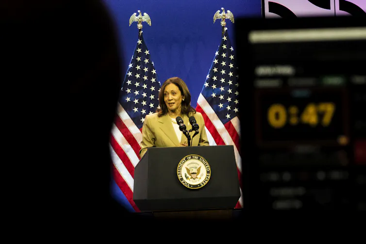 A woman in a beige jacket with dark hair standing at a lectern is photographed from a distance between two structures, one with a clock that reads 47 secconds
