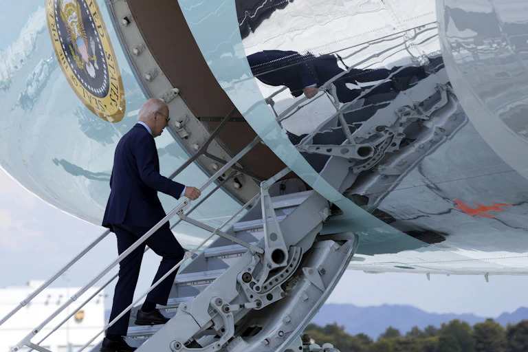 An aging man with thinning grey hair a blue suit walks up the steps to an airliner.