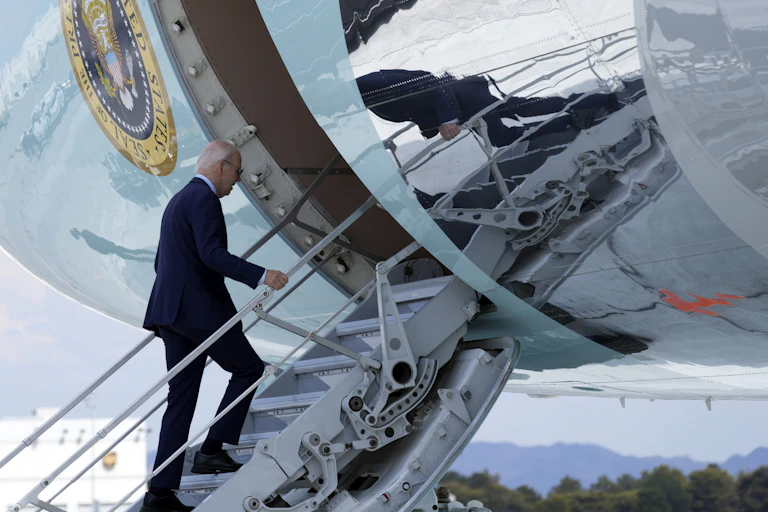 Um homem idoso, com cabelos grisalhos ralos e terno azul, sobe os degraus de um avião.