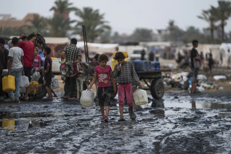 children carrying water bottles walk through muddy area