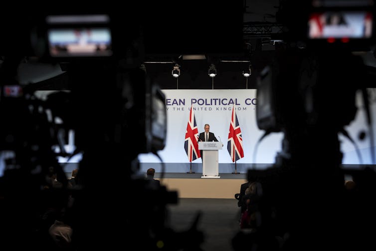 Visão ampla de Starmer falando em um pódio, ladeado por duas bandeiras do Reino Unido. A foto foi tirada por trás de duas câmeras, que são visíveis no primeiro plano.
