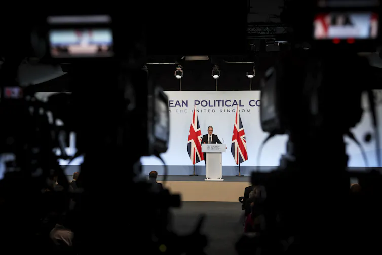 Wide view of Starmer speaking at a podium, flanked by two UK flags. Photo is taken from behind two cameras, which are visible in the foreground.