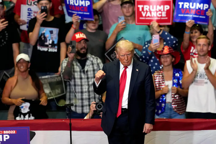 Donald Trump raises a clenched fist as hs stands in front of a crowd of his supporters holding up Trump/Vance placards.