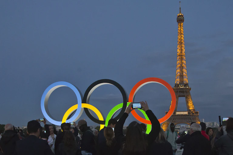 The Olympic Rings lit up with the Eiffel Tower in the background. People in silhouette are taking photos.