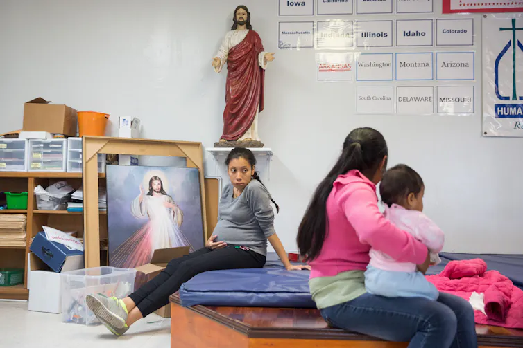 A pregnant woman sits on a bed which has a photograph and a statue of Jesus next to it. Another woman, seated on the adjoining bed, turns her head while holding a child.