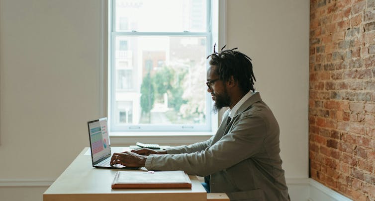A man with a beard and dreadlocks works on a laptop in a home office.