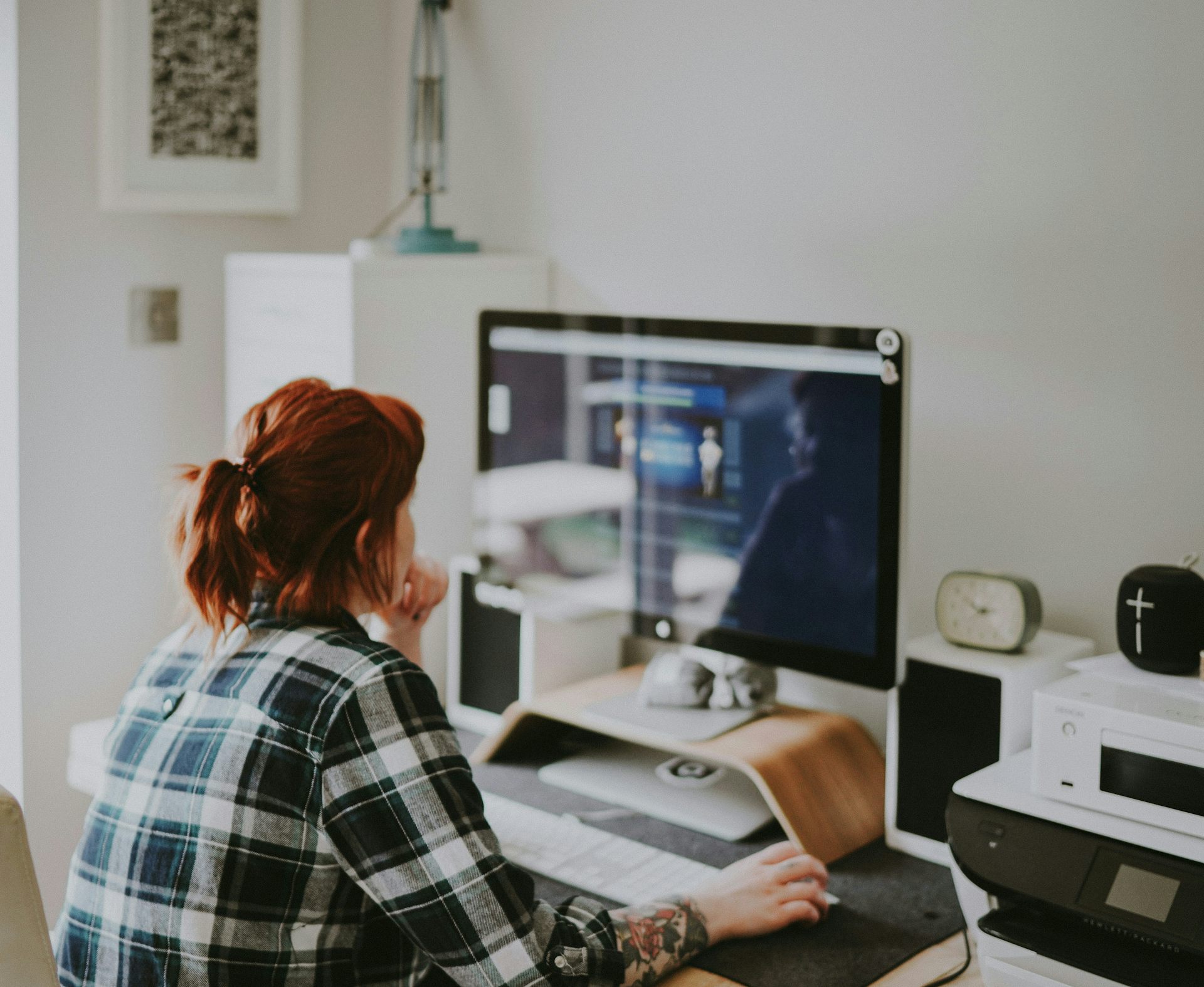 A woman in a plaid flannel shirt sits in front of a computer in a home office.