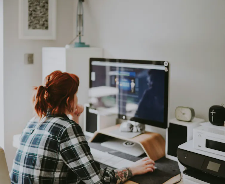 A woman in a plaid flannel shirt sits in front of a computer in a home office.