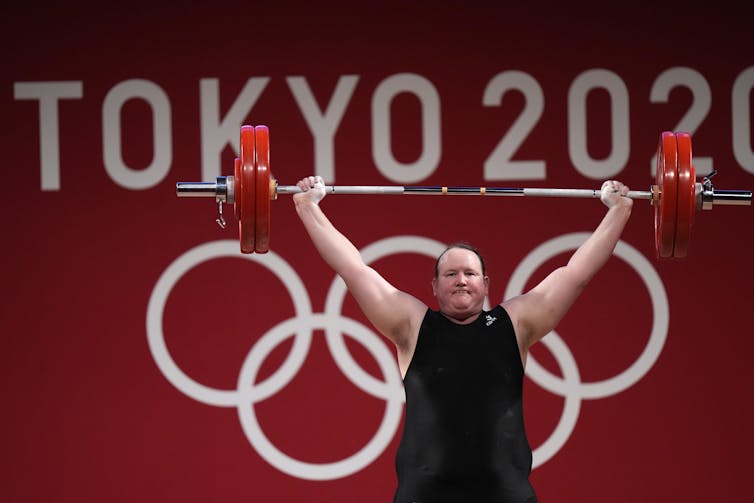 A weightlifter holds a barbell above their head in front of a red wall that says 'Tokyo 2020' on it