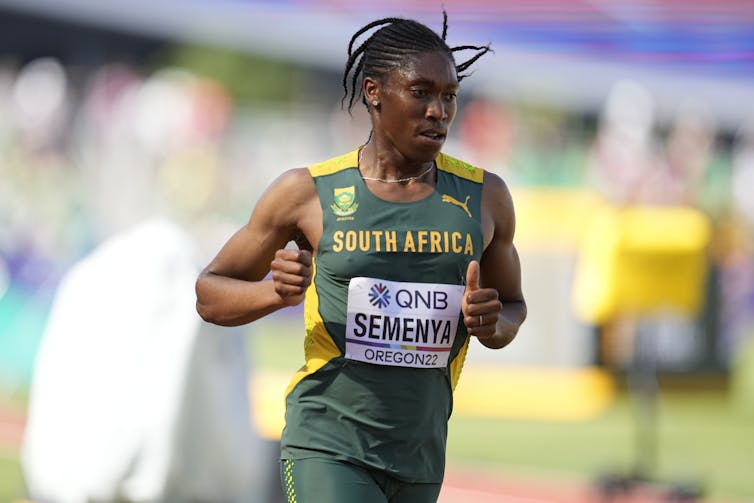 A muscular Black woman in a green and yellow tank top runs a track