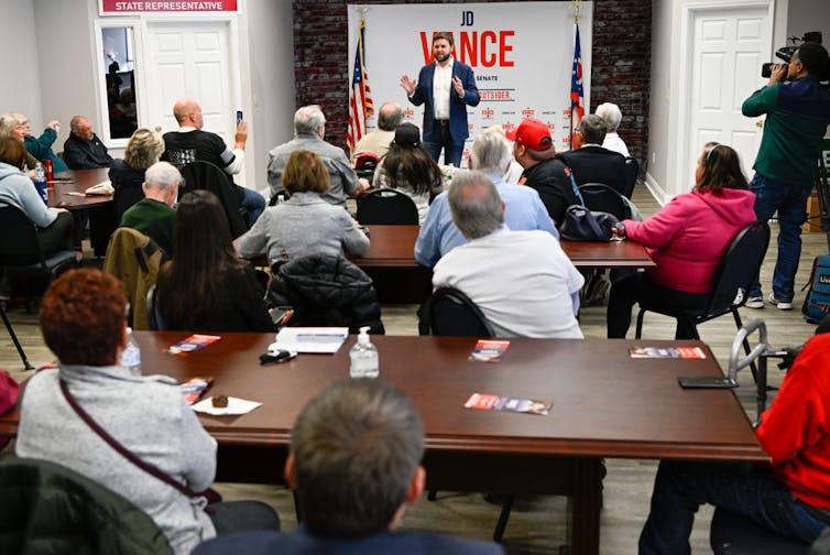 A man with a brown beard and a suit stands and speaks to a room of people seated at tables.
